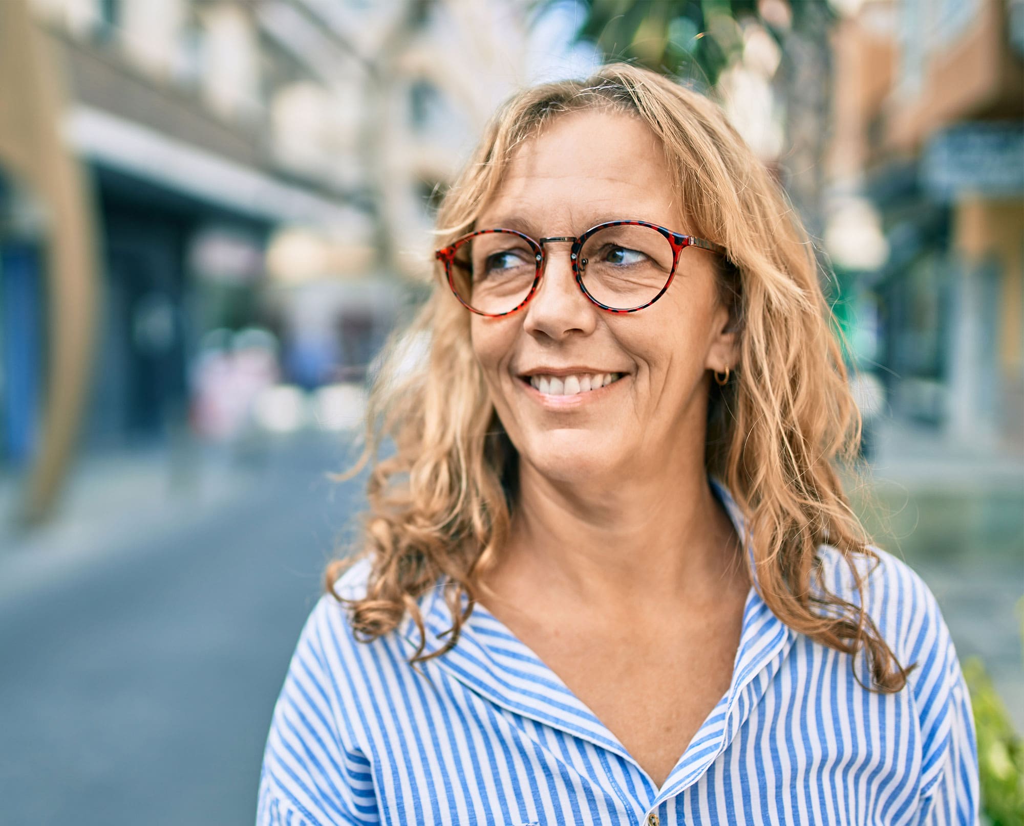 Frau mit brünetten Haaren, gestreifter Bluse und Brille lächelt und sieht nach links – im Hintergrund sieht man städtisches Ambiente